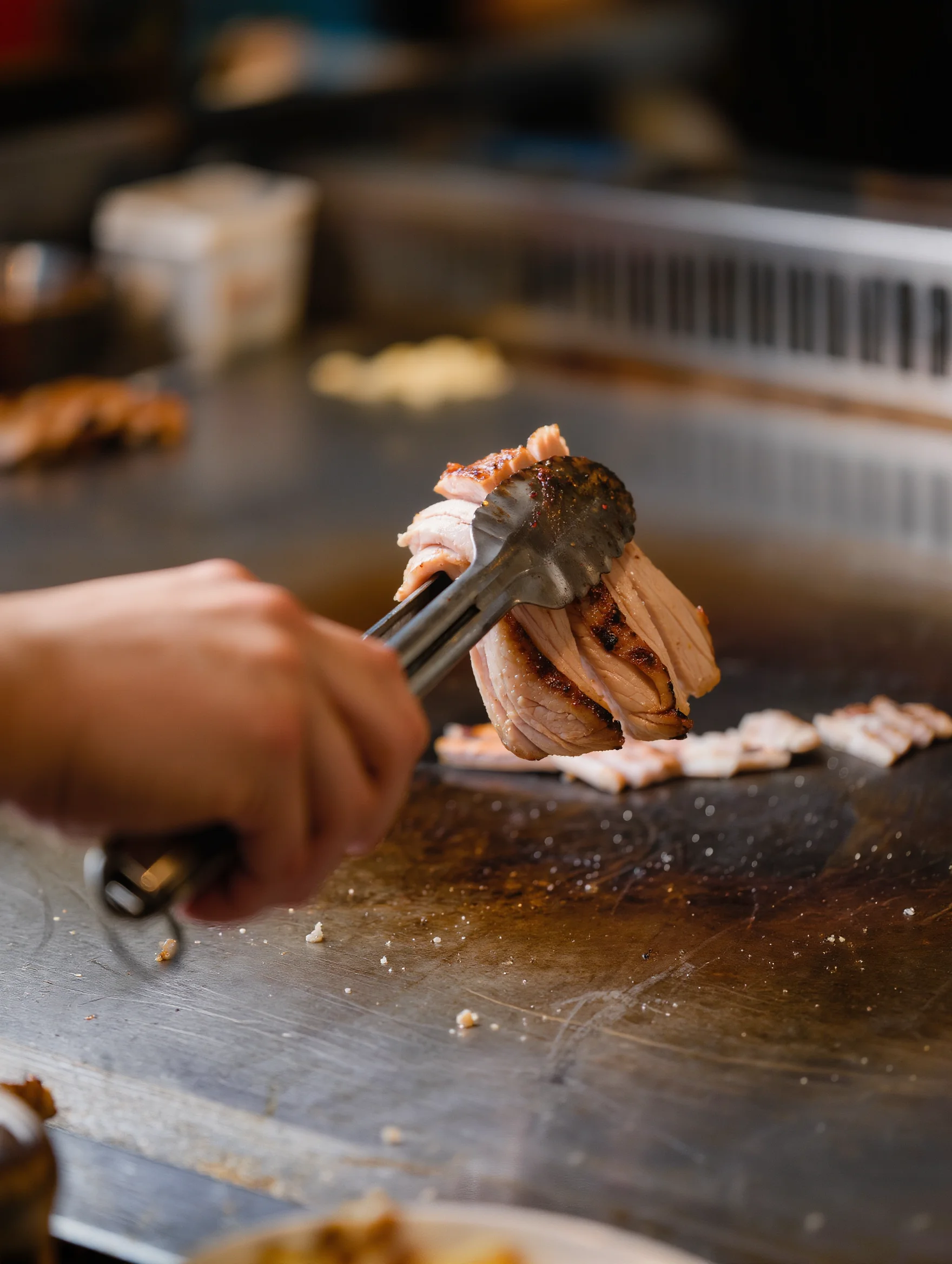 A cook's hand lifting a portion of cooked protein with tongs over a stainless steel prep counter